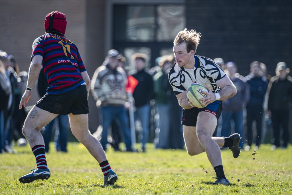 A Westshore player running with the ball past a Castaway Wanderers defender, spectators along the touchline, February 2026.